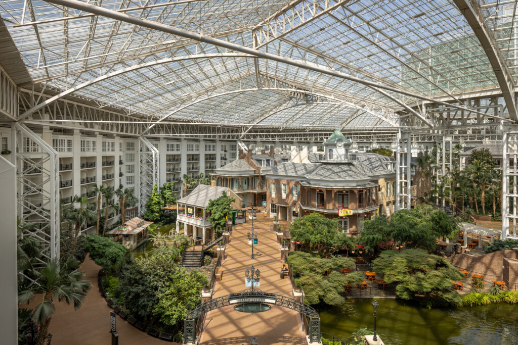 Gaylord Opryland - Expansive glass-roofed atrium featuring replica historic buildings, lush tropical vegetation, a pedestrian plaza with bridge over water, and tall steel framework overhead