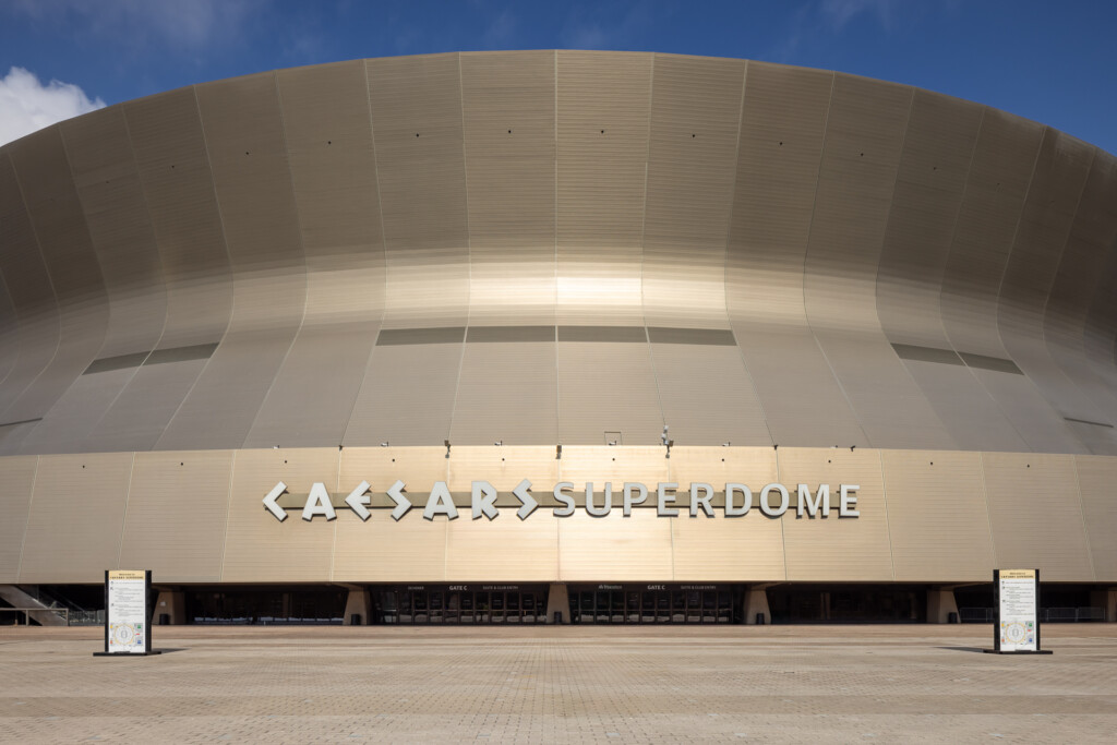 New Orleans Superdome - Large curved beige dome structure with vertical ribbed panels and illuminated signage reading Caesars Superdome, wide plaza in foreground under clear blue sky