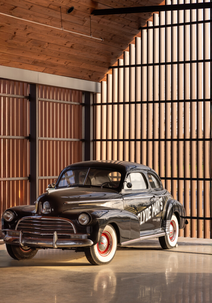 Clyde Mays Distillery - A black 1940s coupe with red-wall tires sits in a concrete garage with exposed wood ceiling and illuminated vertical slat walls.