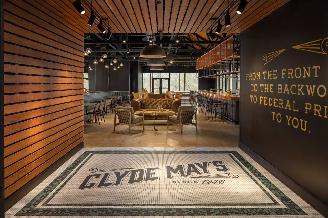 Modern distillery entryway featuring a geometric tile floor with Clyde May's branding, wood-slat accent walls, brass pendant lights, and lounge seating visible beyond the threshold.