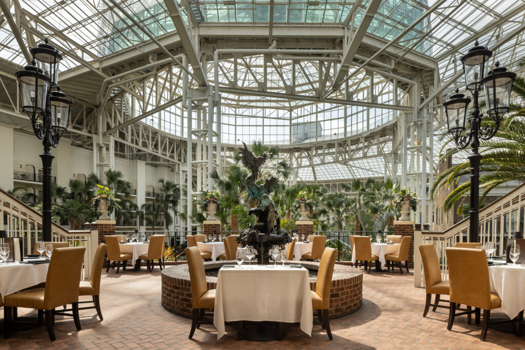 Elegant dining room with round white-clothed tables, tan upholstered chairs, ornamental street lamps, and abundant green plants beneath an expansive glass-paneled roof structure