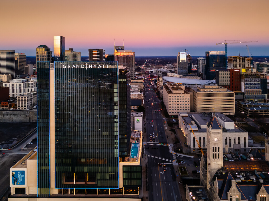 Modern hotel tower with illuminated vertical ribbing at twilight, surrounded by downtown skyline and city streets below