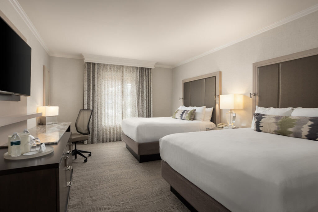 Spacious hotel room featuring two queen beds with white linens, desk with office chair, wall-mounted TV, and large windows with layered curtains