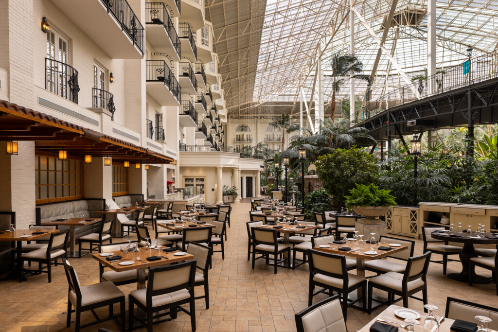 Gaylord Opryland - Modern hotel restaurant with dining tables and chairs beneath a large glass ceiling and steel framework, with a multi-story white building and tropical plants visible