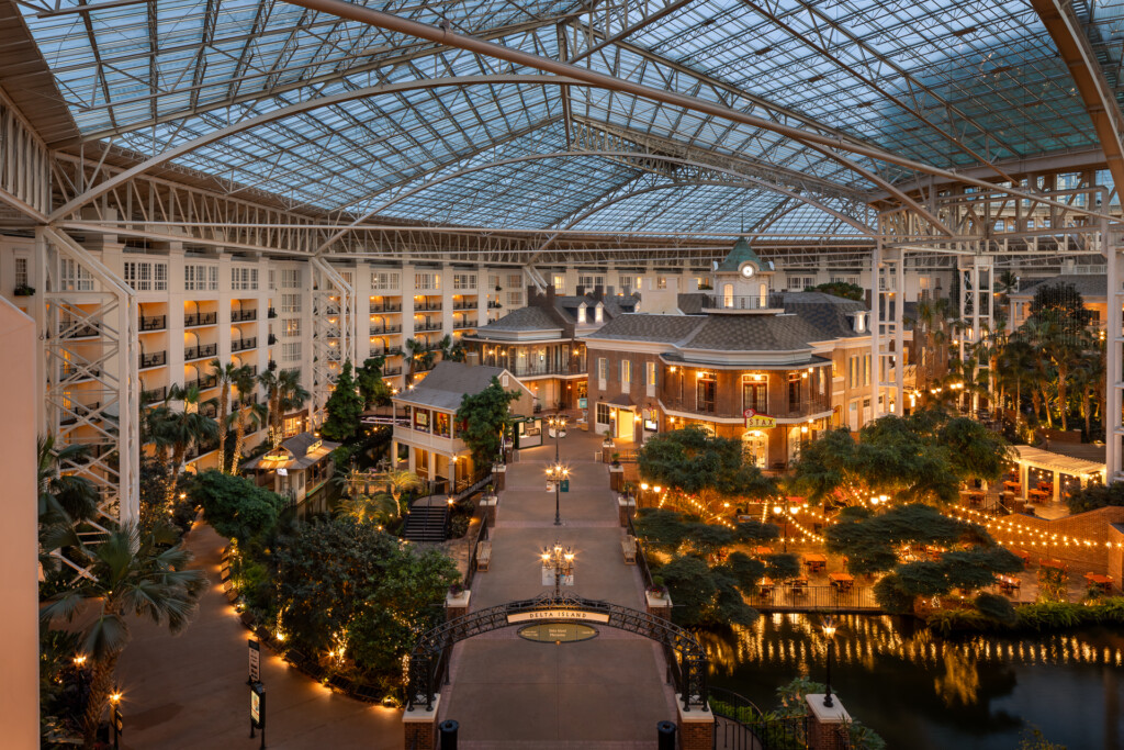 Gaylord Opryland - A sprawling indoor atrium with ornate glass and steel ceiling features colonial-style buildings, lush landscaping, warm lighting, and a water feature at dusk.