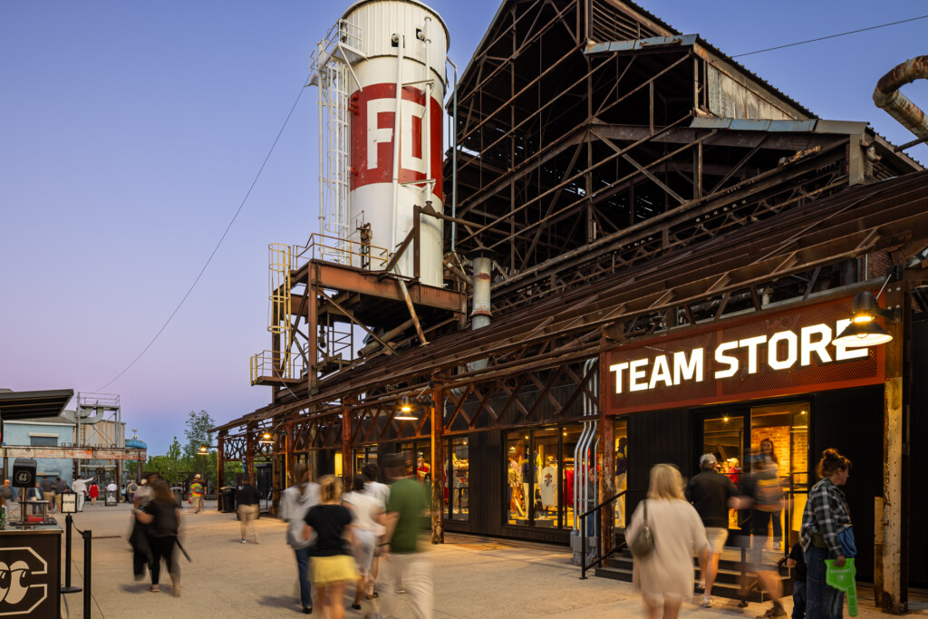 Erlanger Park - Chattanooga Lookouts - Pedestrians walk through an outdoor shopping area with a vintage water tower and steel-frame structures, warm lights glowing from the Team Store storefront.