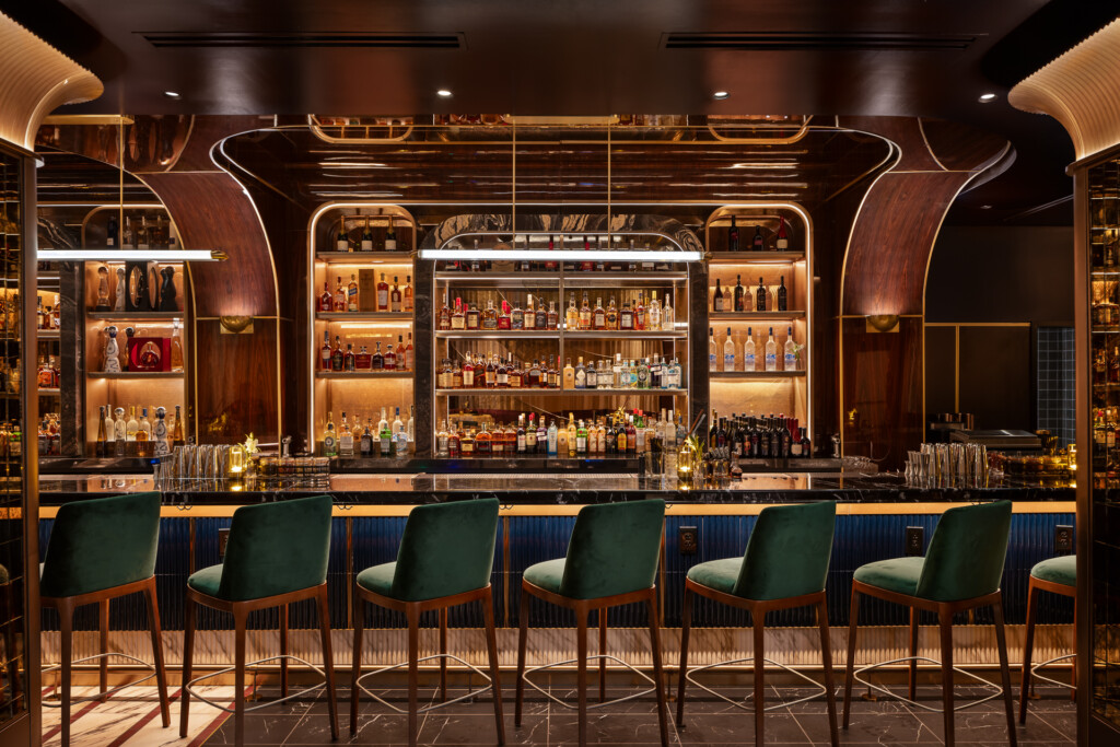 Philippe Chow - Nashville - Dark wood bar with illuminated shelving displaying spirits, green upholstered bar stools, neon accents, and decorative wooden architectural details.