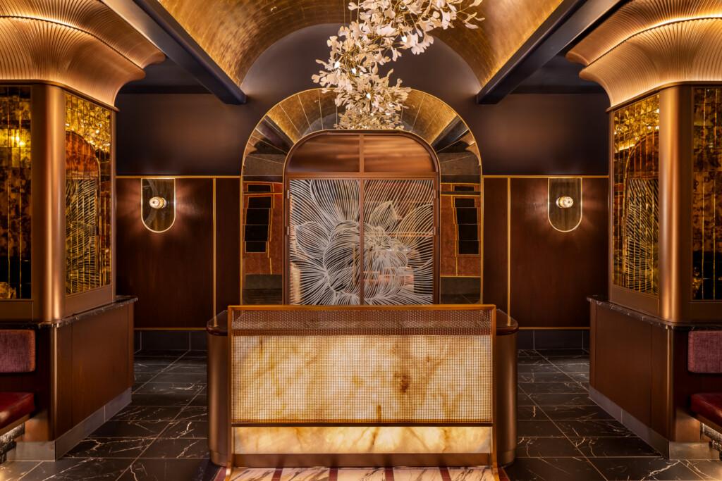 Philippe Chow - Nashville - A symmetrical restaurant lobby features a marble reception desk with brass details, ornate ceiling with curved beams and a floral chandelier, rich brown walls with gold accents, and decorative sconces flanking an artistic floor pattern.
