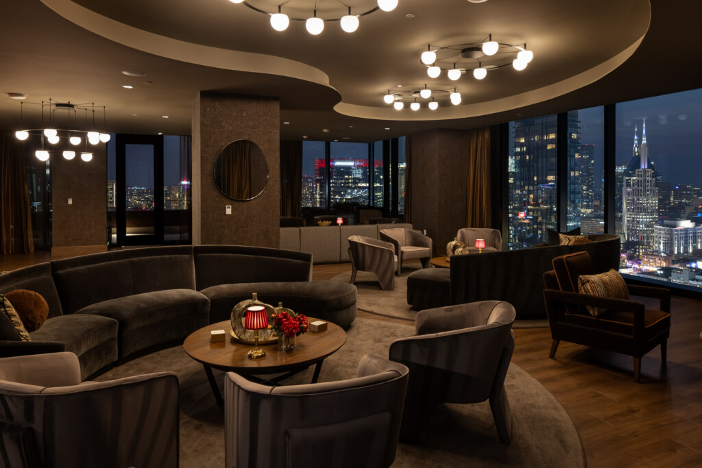 Olive at Peabody - Elegant hospitality lounge with curved charcoal seating, sculptural ceiling details, globe pendant lights, and floor-to-ceiling windows overlooking illuminated city skyline at night