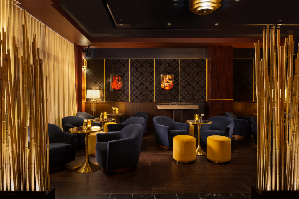 Philippe Chow - Nashville - Sophisticated lounge space with dark upholstered armchairs, gold accent tables, yellow ottomans, warm lighting, and decorative wood paneling framing the room.