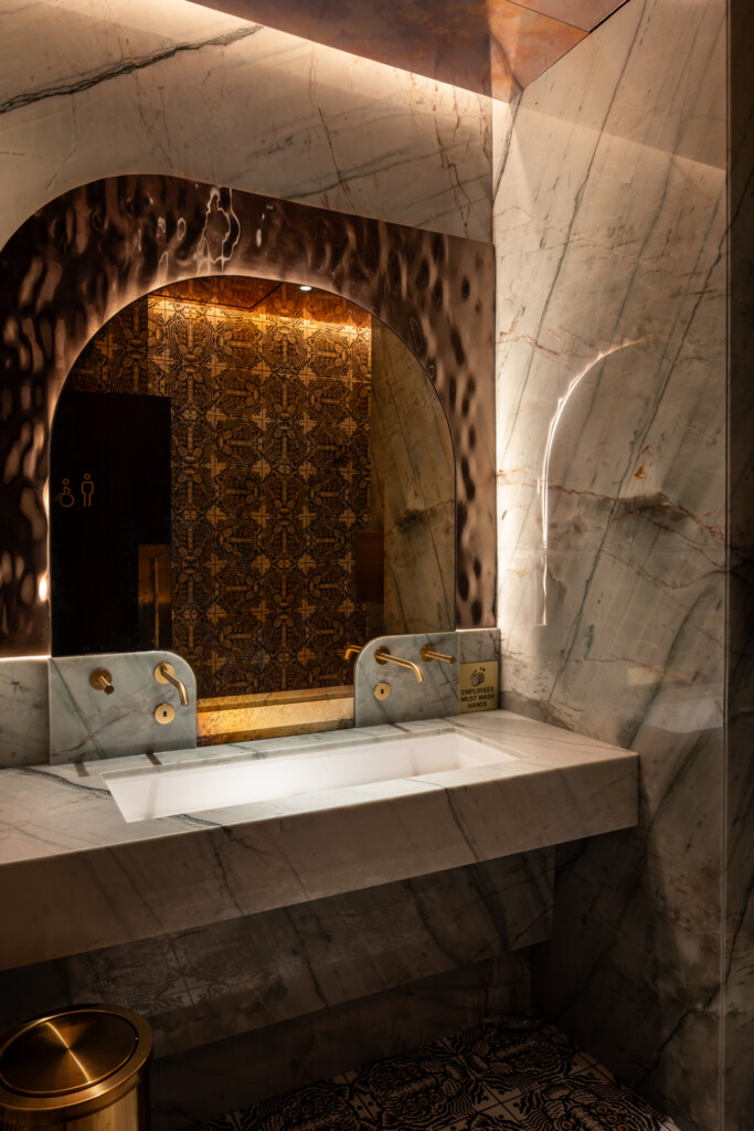 Philippe Chow - Nashville - Marble bathroom with large arched mirror, white sink, brass fixtures, patterned gold wallpaper, and ambient lighting