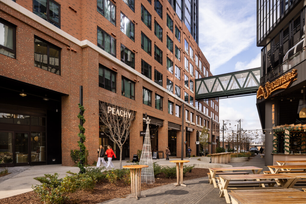 Linea at Southend - Outdoor courtyard space with brick buildings, walkway, wooden picnic tables, pedestrians, and an elevated bridge connecting structures