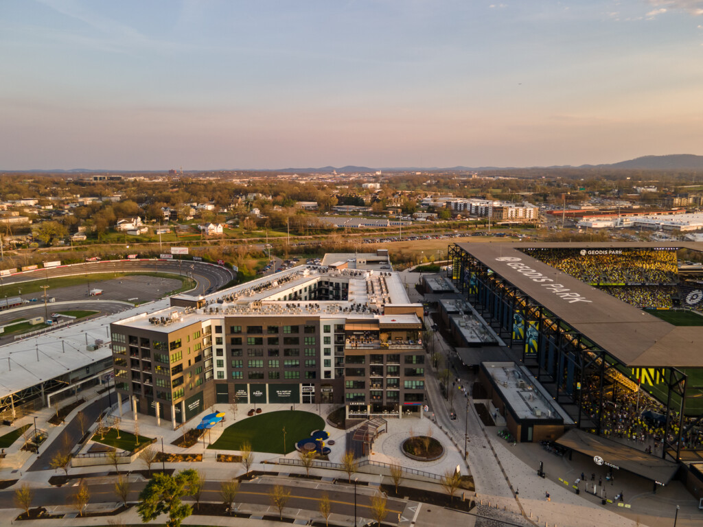 Aerial view of a modern mixed-use building with yellow and white facades, rooftop plaza, surrounding parking lots, sports stadium, and forested residential areas under evening light.