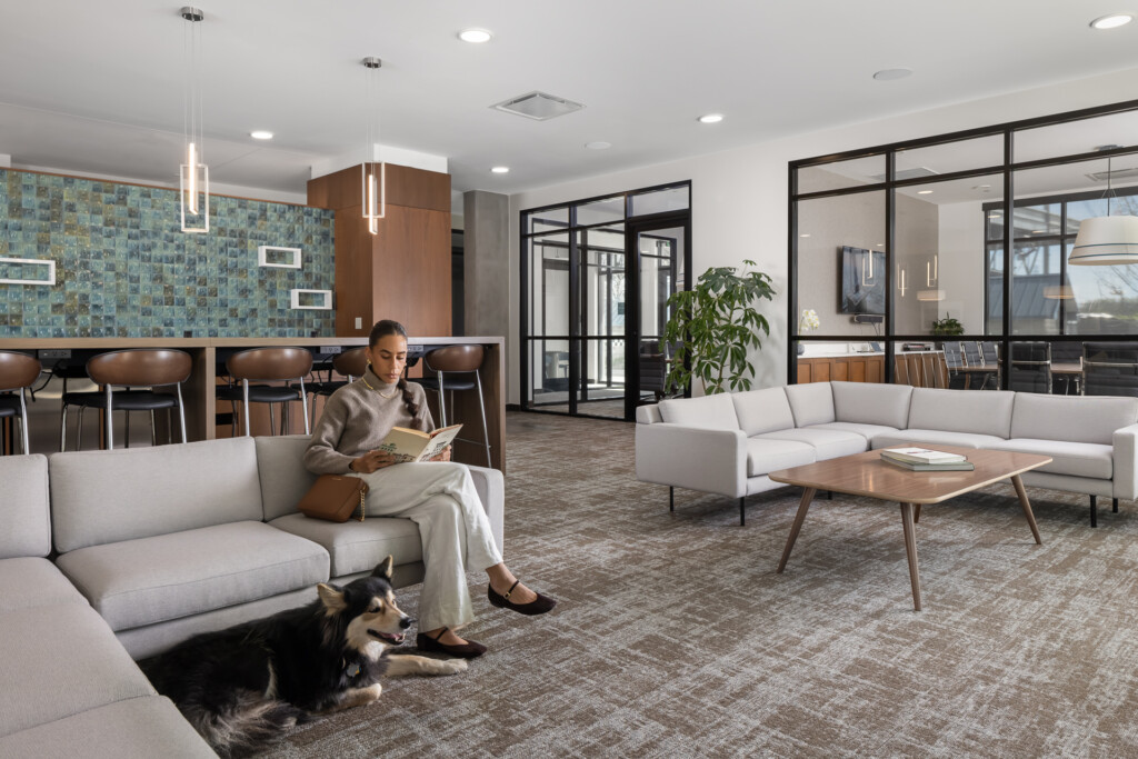 Open-concept interior with light gray modular sofas, wood coffee table, and person reading on sofa with dog. Kitchen bar with turquoise tile backsplash and pendant lights visible in background. Black-framed glass doors and planted areas throughout.