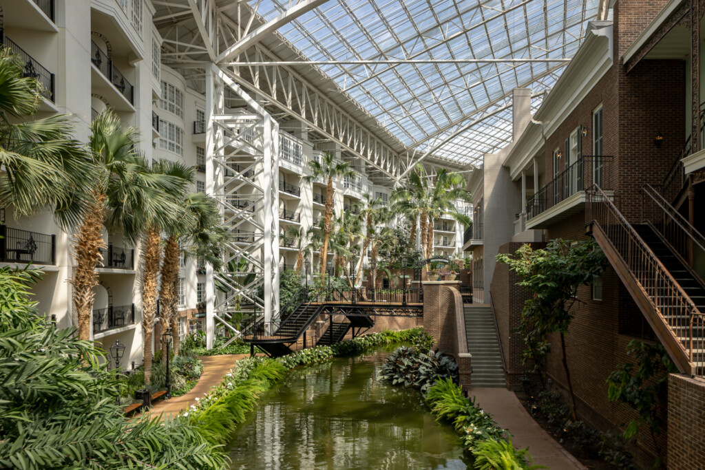 Gaylord Opryland - Interior courtyard with glass ceiling, brick and white residential buildings flanking a reflecting pool lined with palm trees and lush green plants