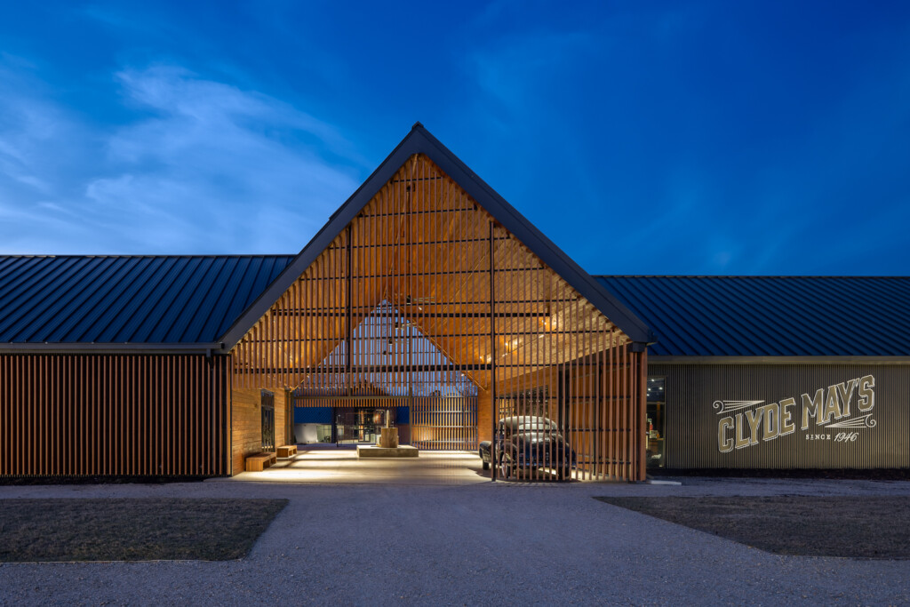 Contemporary barn structure with peaked roof, vertical wood slats, illuminated interior, metal roofing, and gravel courtyard at dusk