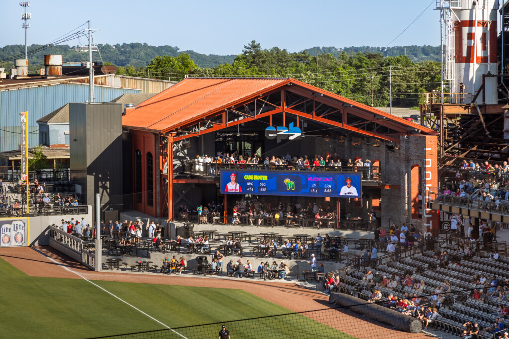 Large outdoor baseball stadium with distinctive orange steel-framed pavilion structure, crowd seating, and manicured grass field in foreground
