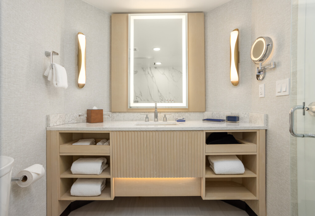 Light wood vanity with open shelving, marble countertop, lighted rectangular mirror, brass sconces, and organized towel storage