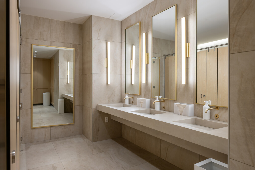Spacious bathroom with long white double vanity, dual brass-framed mirrors, beige tile walls, and glass shower enclosure