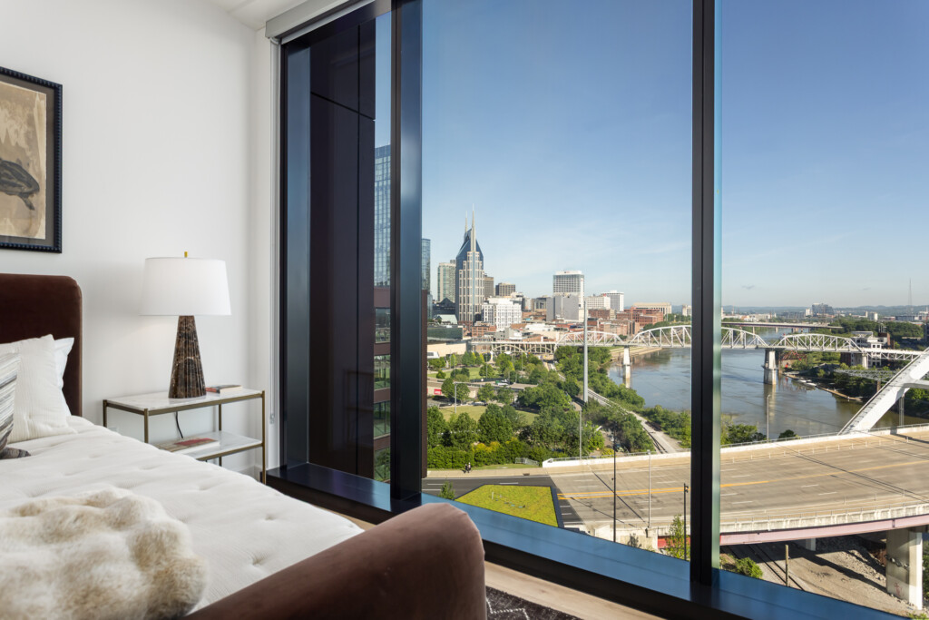 Luxury bedroom with brown upholstered headboard, white bedding, and floor-to-ceiling windows overlooking a downtown skyline, river, and bridge