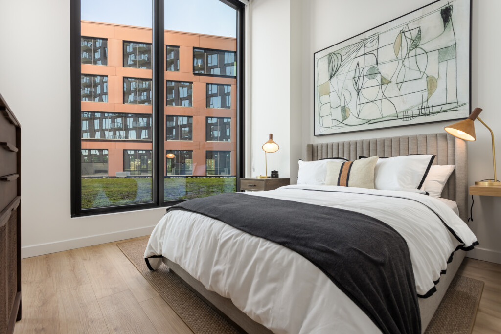Bedroom with upholstered headboard, white bedding with black throw blanket, wooden nightstand, brass lamp, abstract wall art, and large windows overlooking an orange brick building