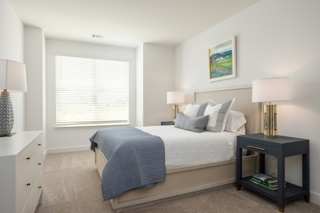 Bright bedroom featuring a beige upholstered bed with blue throw blanket, white nightstands, black side table, table lamps, and large window with plantation shutters