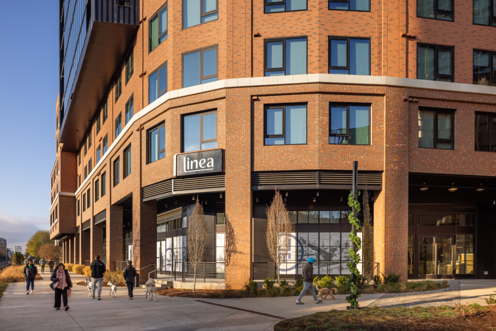 Curved brick building with large windows and ground-level retail space, pedestrians walking on plaza