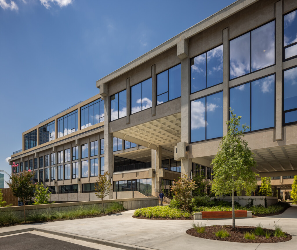 Contemporary multi-story commercial building with large gridded windows, concrete structural elements, and landscaped plaza with young trees and plantings