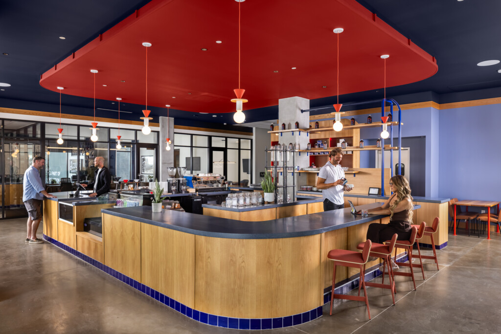 Open-concept kitchen with curved wooden service counter, red suspended ceiling, pendant lighting, and seating area with guests and staff interacting.