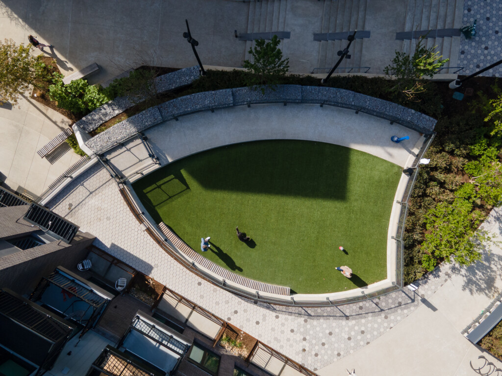 Overhead view of a curved courtyard with lush green lawn, concrete seating areas, metal benches, and people relaxing on the grass surrounded by building walls and landscaping