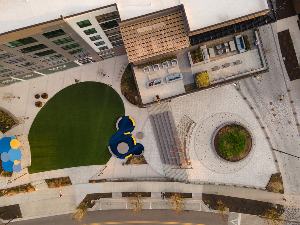 Aerial view of a contemporary courtyard featuring a large green lawn, blue sculptural seating elements, paved walkways, and multi-story residential or commercial buildings with modern design