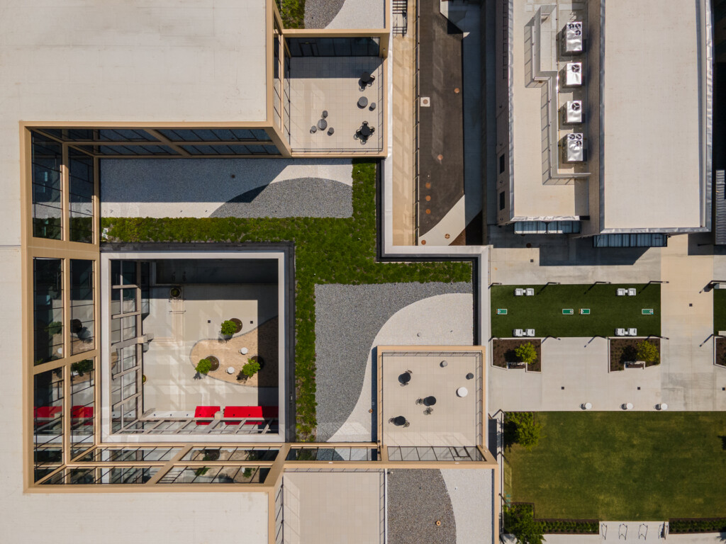 Aerial view of a contemporary courtyard with wooden pergola, red seating, green planted areas, paved pathways, and surrounding residential buildings