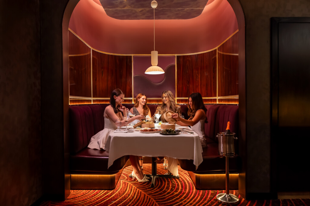 Philippe Chow - Nashville - Four women seated at a round table with white tablecloth in a contemporary dining space with curved burgundy banquettes, gold-trimmed walls, pendant lighting, and patterned orange carpet