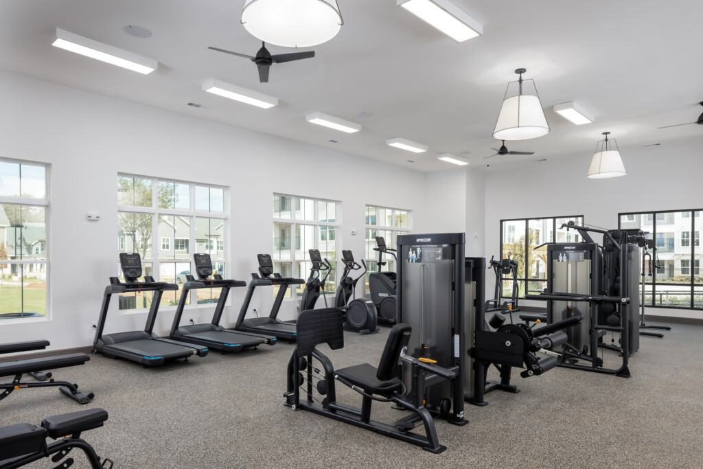 Bright fitness center with rows of treadmills, exercise machines, pendant lighting, and large windows overlooking landscaped grounds