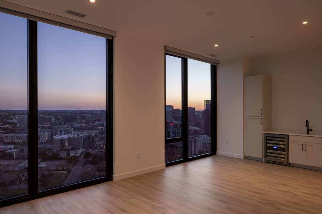 Empty residential interior with floor-to-ceiling windows overlooking cityscape at dusk, wooden floors, recessed lighting, and a wet bar with wine cooler