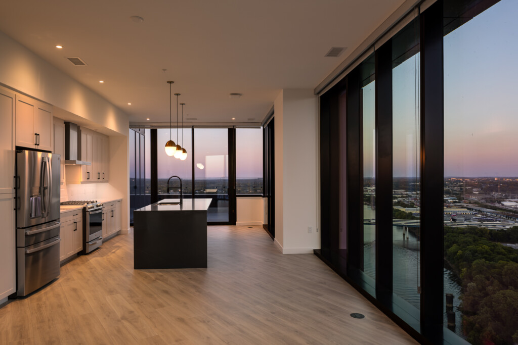 Open-concept kitchen with stainless steel appliances and black island, expansive windows overlooking cityscape at dusk with pendant lighting above counter.
