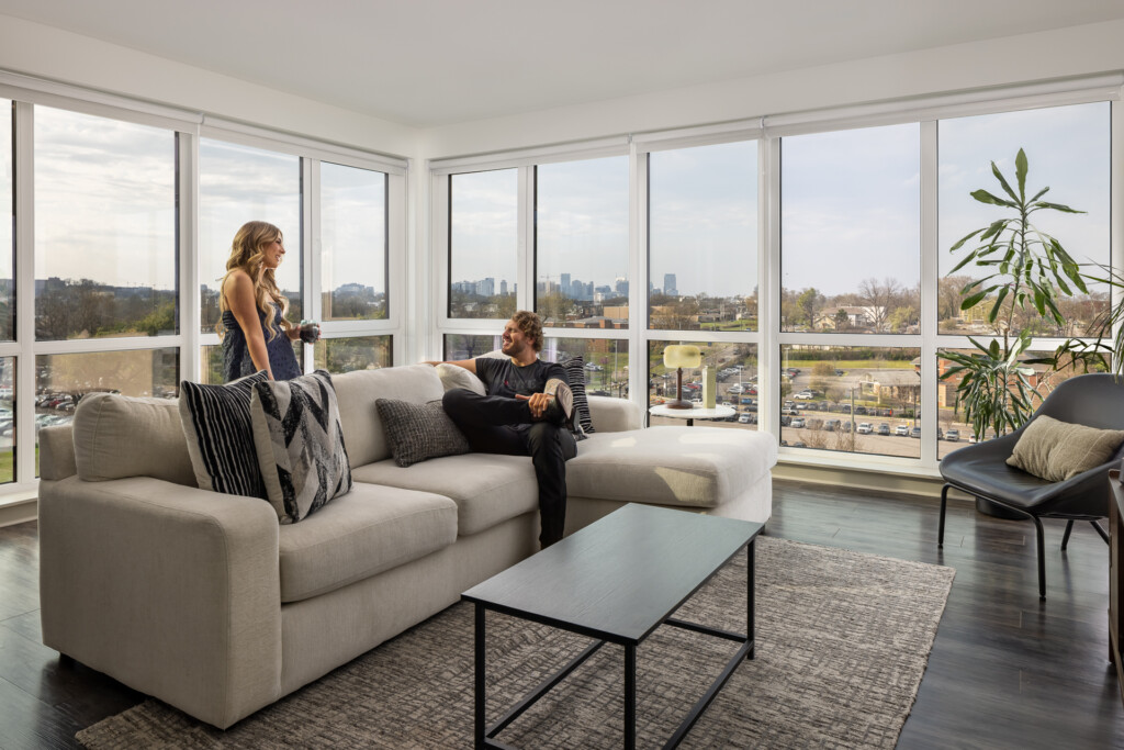 445 Park Commons - GEODIS - Two people seated on a cream sectional sofa in a bright living room with floor-to-ceiling windows overlooking a cityscape, featuring a black coffee table and potted plant