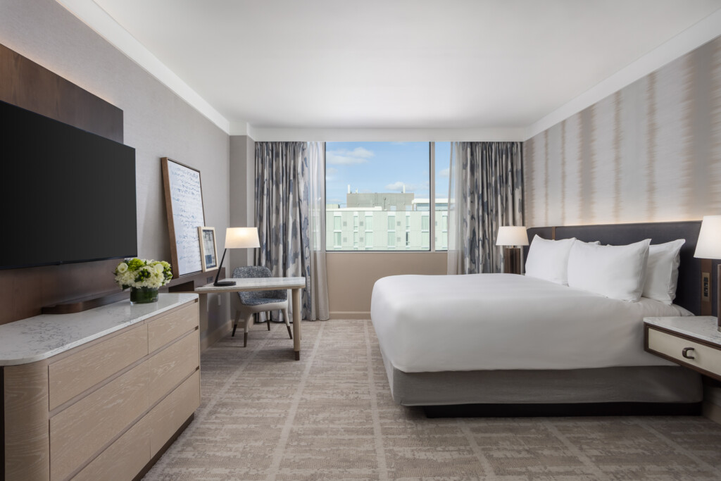 Loews Vanderbilt - Bright hotel room featuring a king bed with white linens, wood dresser with flat-screen TV, work desk, and floor-to-ceiling windows with gray curtains overlooking a cityscape.