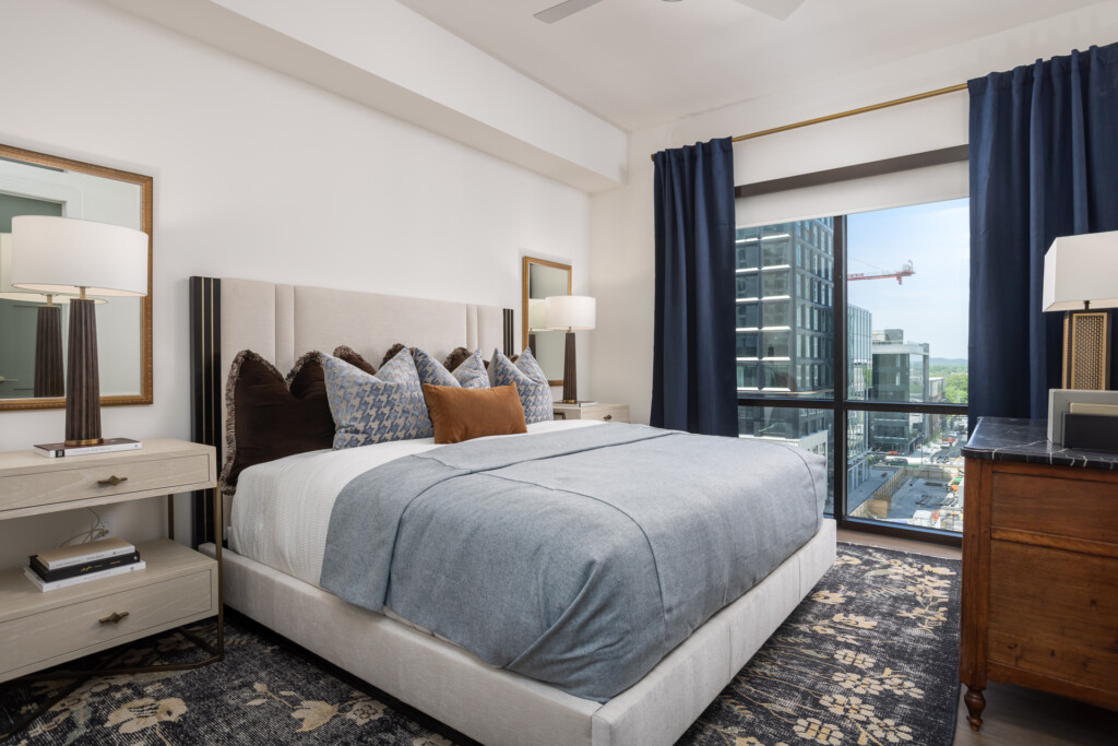 Spacious bedroom with upholstered bed, navy curtains, wooden nightstands, table lamps, and floor-to-ceiling windows overlooking a cityscape