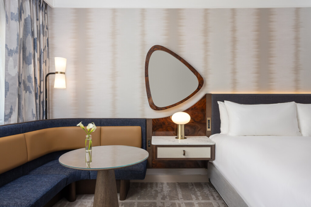 Loews Vanderbilt - Contemporary hotel bedroom with a bed, curved wood-framed mirror, blue banquette seating with tan cushions, round side table, and brass task lighting.