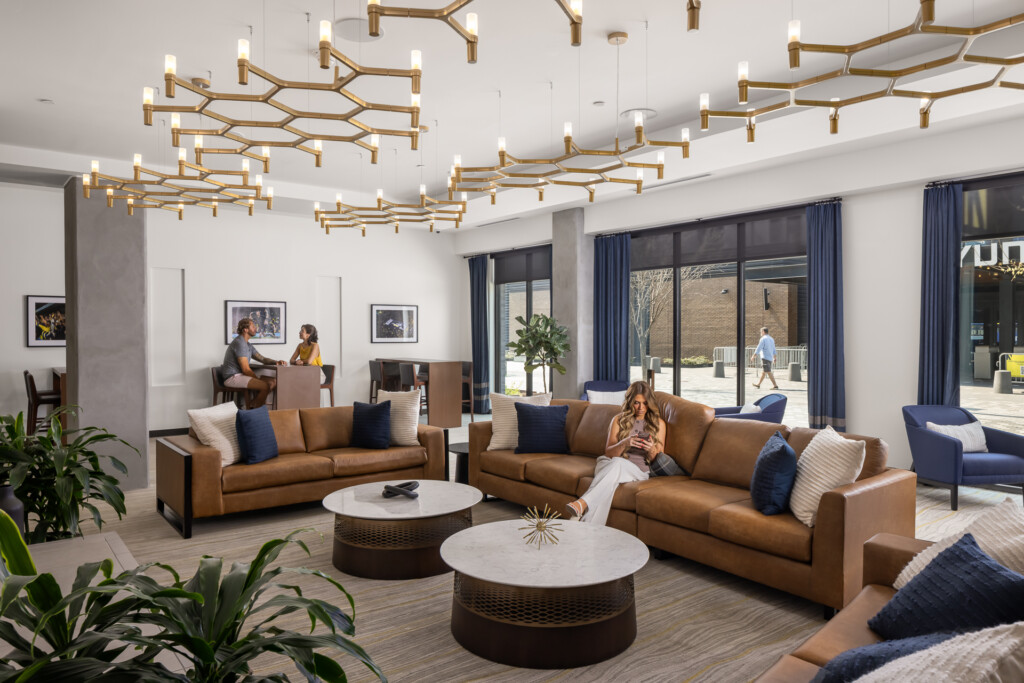 445 Park Commons - GEODIS - Spacious hotel lobby with brown leather sofas, white marble tables, geometric brass branch chandeliers, large windows overlooking outdoor patio, and blue accent furnishings throughout