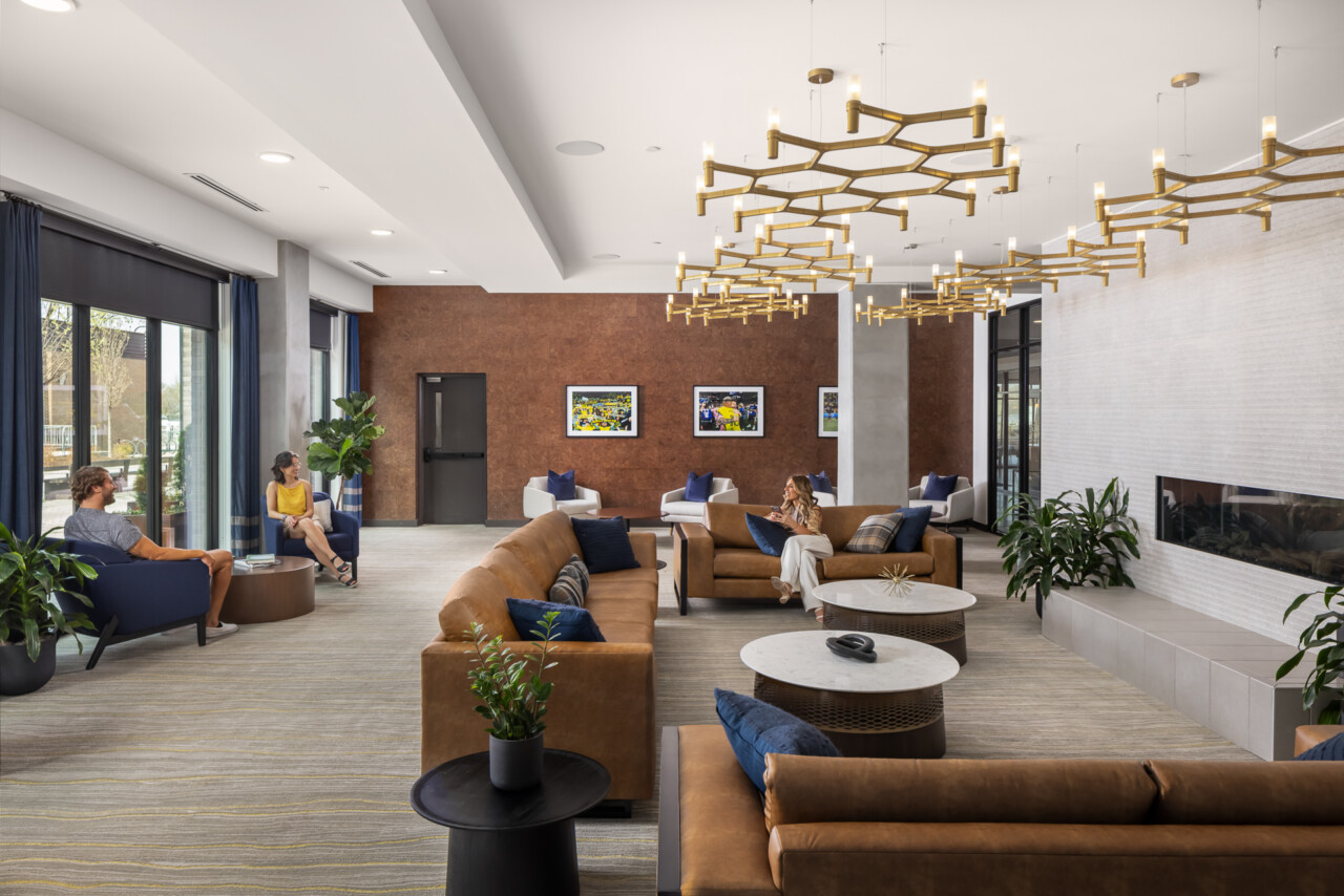 445 Park Commons - GEODIS - Spacious hotel lobby with tan leather seating, brass branch-style pendant lights, cork accent wall, white ceiling, and guests gathered in conversation areas.