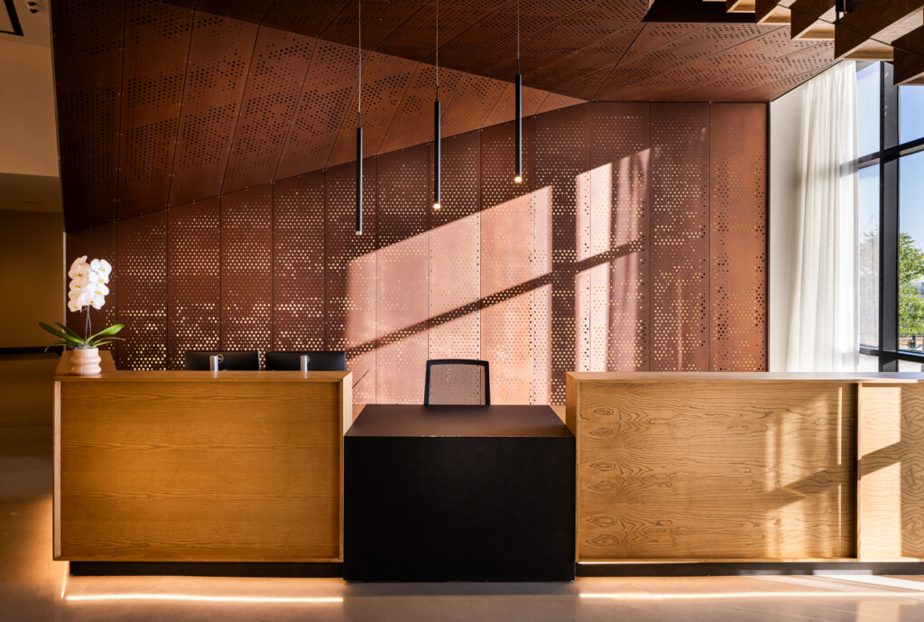 Contemporary hotel lobby featuring two wooden reception desks flanking a central black desk, with a perforated copper screen wall, pendant lights, and bright natural light streaming through large windows