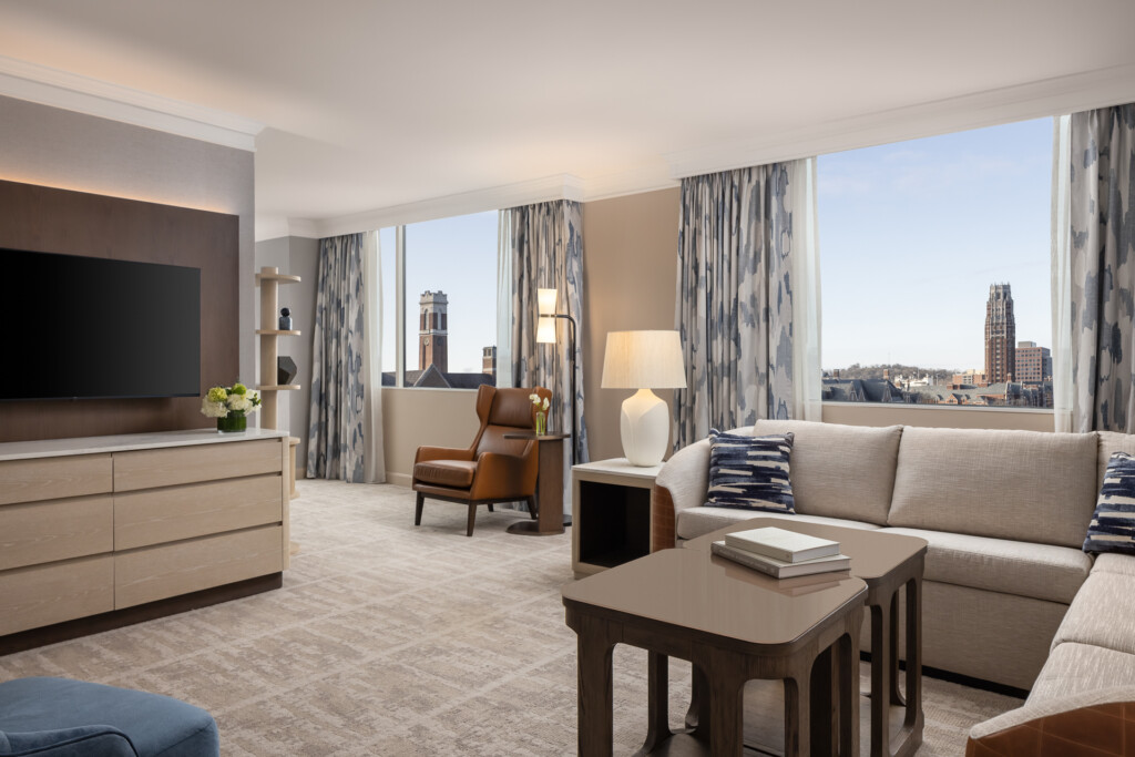Loews Vanderbilt - Spacious hotel suite featuring a beige sectional sofa, brown leather armchair, light wood furniture, and floor-to-ceiling windows overlooking downtown buildings