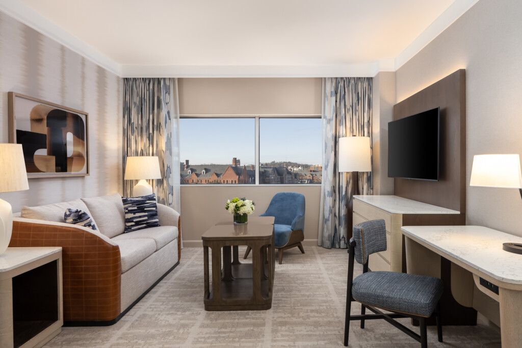 Loews Vanderbilt - Bright hotel suite with curved sofa, blue accent chair, wood table, flat-screen TV, and large windows overlooking cityscape