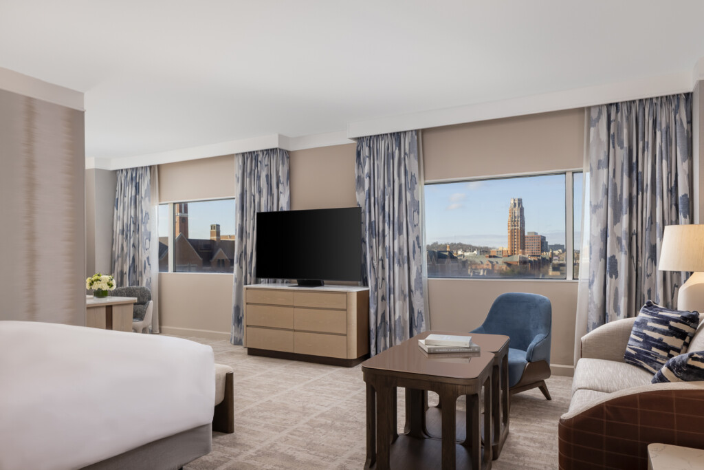 Loews Vanderbilt - Spacious hotel bedroom with king bed, living area with sofa and armchair, flat-screen TV on wood dresser, floor-to-ceiling windows overlooking cityscape, blue patterned curtains and neutral tones throughout
