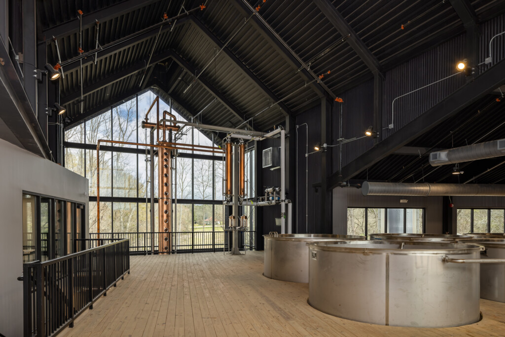 Clyde Mays Distillery - Large metal fermentation tanks in a brewery space with black metal railings, dramatic vaulted corrugated ceiling, and expansive windows overlooking bare trees.