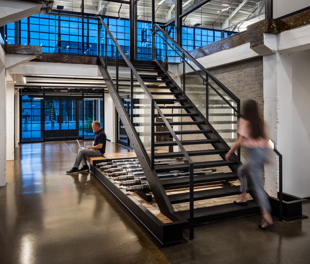 Black steel staircase with wood treads in a bright loft with large windows, exposed beams, and polished concrete floors. Two people move through the space.