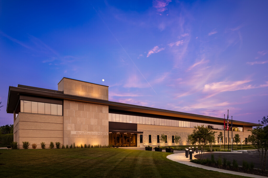 Contemporary institutional building with tan brick and metal cladding, illuminated at twilight, featuring a curved entry plaza with landscaping and flags