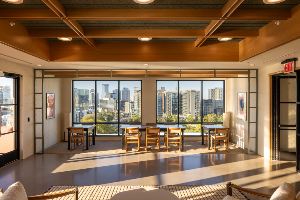 Bright commercial lounge with warm wood ceiling beams, floor-to-ceiling windows overlooking city skyline, wooden chairs, polished concrete flooring, and recessed lighting.